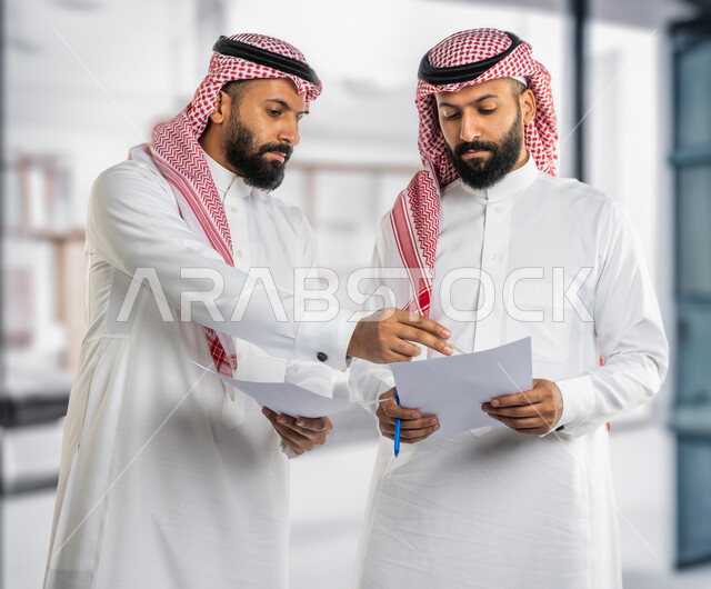 تعزيز الروح التشاركية، توزيع المهام المطلوبة لتطوير أهداف الشركة ، رجلان توأم عربيان خليجيان سعوديان يرتديان الزي التقليدي و يحملان اوراق عمل، اجتماع الزملاء للمناقشة في حل المشكلات، الوحده والعمل الجماعي، بيئة عمل سعودية