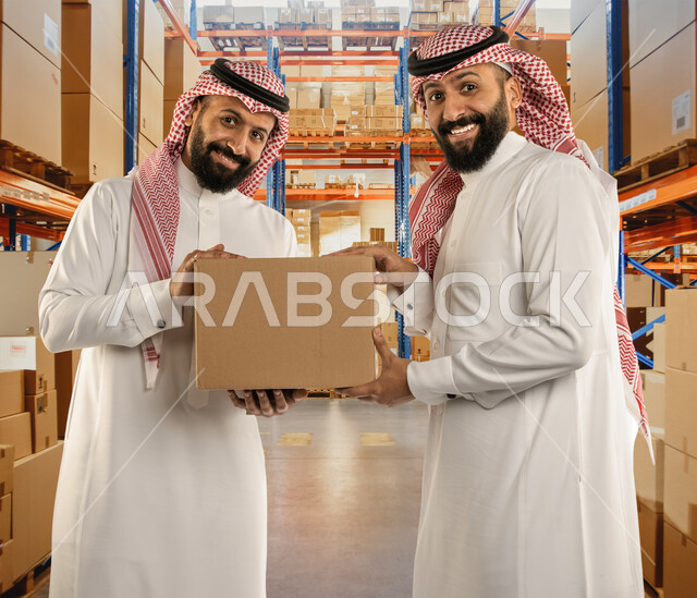 تحضير طلبيات جديدة, رجلان توأم عربيان خليجيان سعوديان يحملان صندوق كرتوني ويقفان في موقع العمل بإيماءات السرور، صناديق كرتون تعبئة وتغليف، بضائع عربية سعودية خليجية،  خلفية صناديق وكراتين مغلفة.