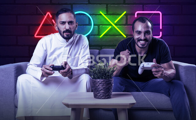 The concept of entertainment, entertainment, victory and victory, two Saudi Gulf Arab men sitting in the living room holding a joystick, enjoying playing video games in their free time, challenge, adventure and competition, advanced modern technologies, a background of illuminated symbols for PlayStation games on a stone wall