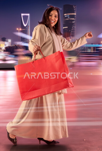 ايماءات السعادة بعد الشراء وتأثيره الإيجابي على المرأة, الاستفادة من العروض الموسمية والتخفيضات, سيده عربيه سعودية خليجية مبتسمة ترتدي العباءه و تحمل بيدها أكياس التسوق، زيارة المولات التجارية ، صورة الجسم بالطول الكامل، خلفية الابراج المضيئة ليلا في الرياض