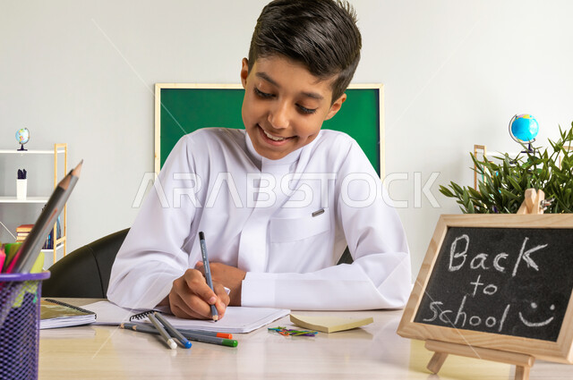 تأدية الواجبات المدرسية، سبورة صغيرة مكتوب عليها نص العودة إلى المدارس، صورة مقربه لطالب عربي خليجي سعودي يرتدي الزي التقليدي يجلس على الطاولة بجانبه مجموعة من الأقلام والكتب المدرسية، الإستعداد للعام الدراسي الجديد بايماءات السرور، التعليم في السعودية