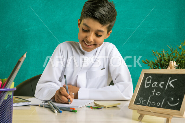 سبورة صغيرة مكتوب عليها نص العودة إلى المدارس، صورة مقربه لطالب عربي خليجي سعودي يرتدي الزي التقليدي يجلس على الطاولة بجانبه مجموعة من الأقلام والكتب المدرسية، تأدية الواجبات المدرسية، الإستعداد للعام الدراسي الجديد بايماءات السرور، التعليم في السعودية