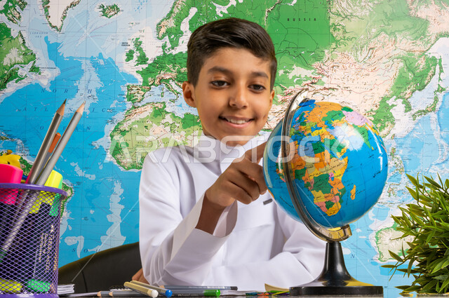The concept of research and exploration around the world, a close-up image of a Saudi Gulf Arab student wearing traditional clothing sitting at the table looking at the globe with a group of pens next to him, preparing for the new academic year, education in Saudi Arabia, returning to school, world map background