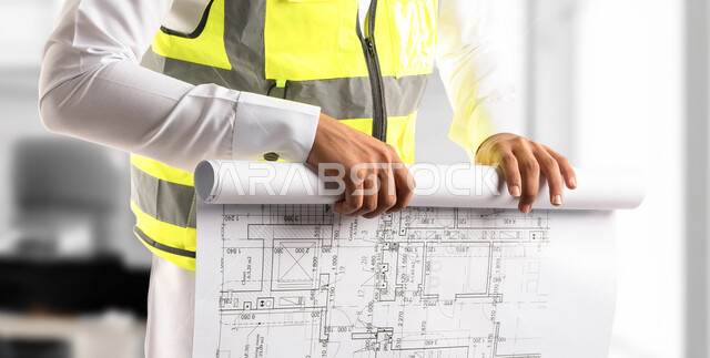 Following up on the project and checking the plans, a close-up picture of a Saudi Arabian Gulf architect wearing a work protective jacket holding the construction plan in his hand, supervision and control of engineering projects, the concept of engineering and architectural construction.