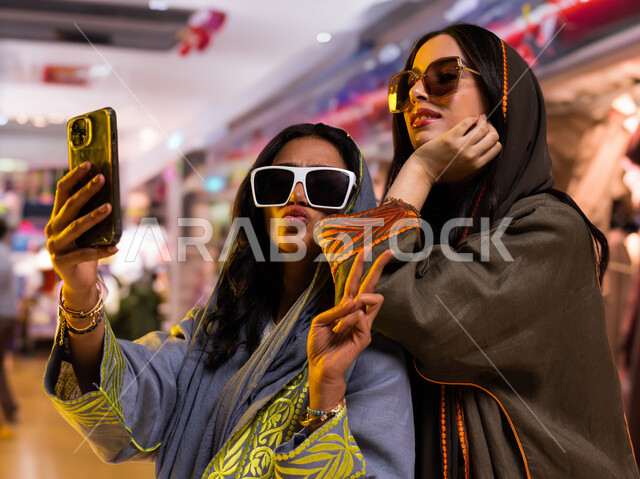إيماءات أنوثة شبابية ظريفة، مفهوم التصوير والشهرة عبر برامج التواصل الاجتماعي، التقاط صورة سلفي عبر الجوال، صورة مقربة لرفيقتان عربيتان خليجيتان سعوديتان ترتديان عباءات خليجية عصرية ونظارات شمسية، قضاء أوقات ممتعة مع الأصدقاء، خلفية محلات تجارية