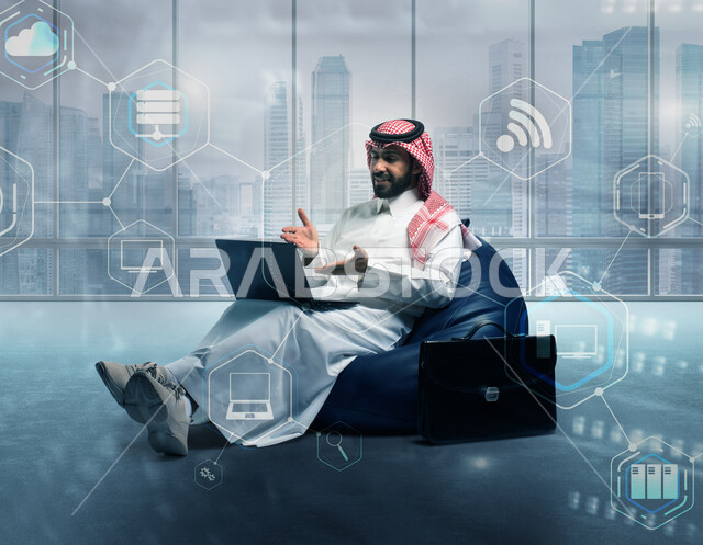 العمل عن بعد باستخدام اللابتوب ، رجل أعمال عربي سعودي خليجي يرتدي الشماغ و الثوب السعودي يجلس على كرسي مريح بجانبه حقيبة ويقوم بإجراء مكالمة فيديو باستخدام الحاسوب المحمول, إيماءات تدل على شرح وتوضيح الأفكار, خلفية أيقونات تقنية هولوجرامية مختلفة