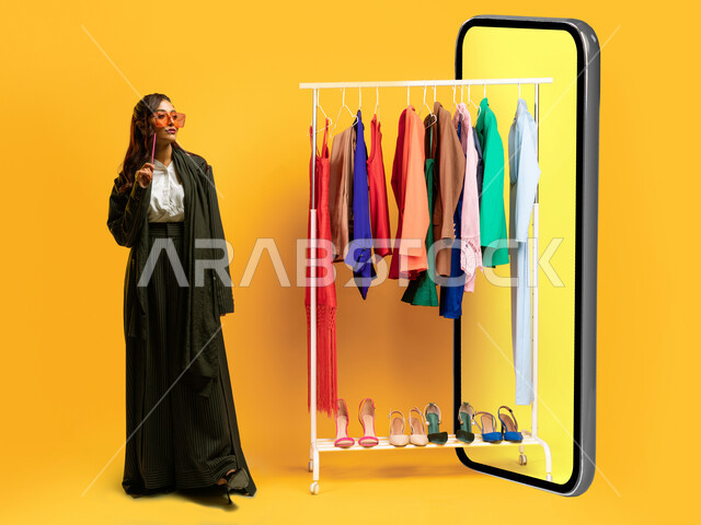 مجسم لموبايل كبير الحجم بشاشة صفراء تخرج منها علاقة ملابس, التسوق وشراء الألبسة عبر الانترنت، بورتريه لسيدة أعمال عربية خليجية سعودية ترتدي اللباس الرسمي تمسك بيدها قلم و تنظر إلى شيء ما, الاهتمام والاعتناء بالمظهر الخارجي, صورة الجسم بالطوال الكامل، خلفية صفراء