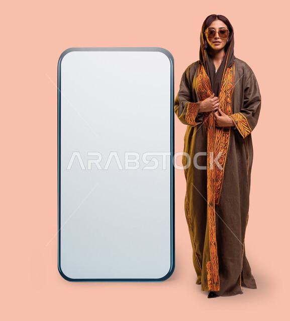 إيماءات الأنوثة والجمال, عرض الأزياء بالتصاميم والموديلات الحديثة، بورتريه لامرأة عربية خليجية سعودية ترتدي عباءة بنية مطرزة ونظارات شمسية عصرية تقف بثقة بجانب مجسم لهاتف محمول كبير بشاشة بيضاء فارغة، موك أب لمربع أبيض كبير، صورة الجسم بالطول الكامل، خلفية وردية