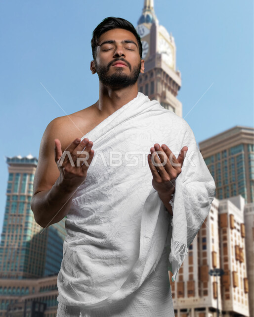 القيام بفريضة الحج و زيارة بيت الله الحرام, إيماءات وجه تدل على الخشوع الى الله, أداء مناسك العمرة و التقرب الى الله في شهر رمضان, صورة مقربة لشاب عربي سعودي خليجي يرتدي ملابس الإحرام و يرفع يديه للدعاء والاستغفار، خلفية برج الساعة و أبراج الحرم المكي