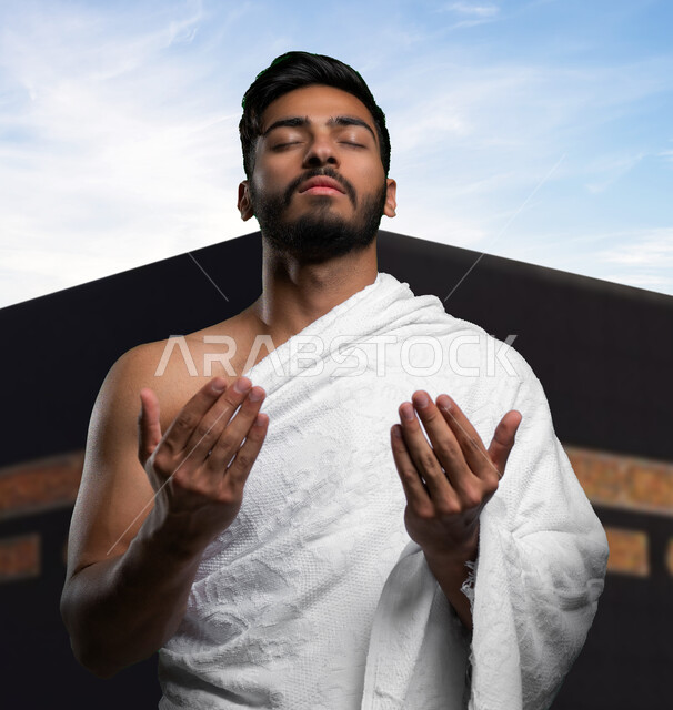 إيماءات وجه تدل على الخشوع الى الله, أداء مناسك العمرة و التقرب الى الله في شهر رمضان, صورة مقربة لشاب عربي سعودي خليجي يرتدي ملابس الإحرام و يرفع يديه للدعاء والاستغفار، القيام بفريضة الحج و زيارة بيت الله الحرام, خلفية الكعبة المشرفة