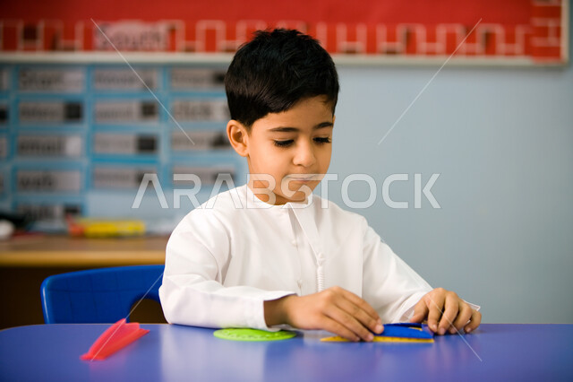 التعلم عن طريق اللعب داخل الفصل، استخدام الأشكال الهندسية والتعرف على أشكالها، صورة مقربة لطالب عربي خليجي إماراتي يرتدي الزي الاماراتي ويمسك البوصلة, الاستمتاع بالنشاطات الترفيهيه لتلاميذ التمهيدي