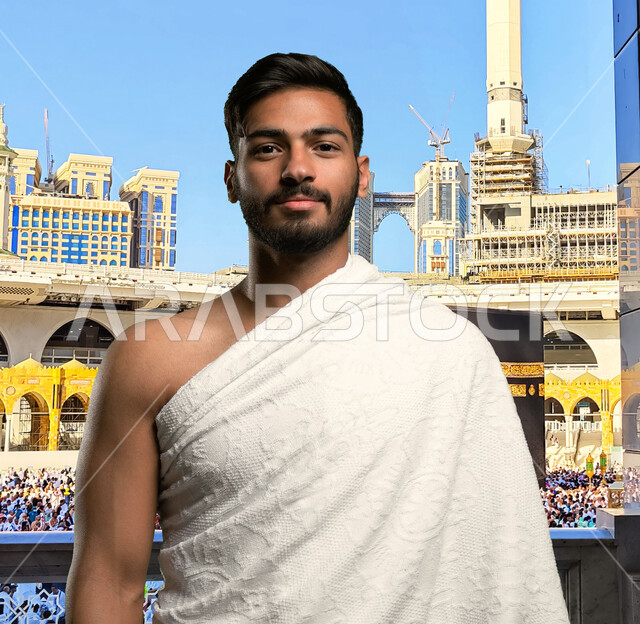 أداء العمرة و التقرب الى الله في شهر رمضان, صورة مقربة لشاب عربي سعودي خليجي مبتسم يرتدي ملابس الإحرام و يستعد لأداء مناسك الحج والعمرة، إيماءات وجه تدل على السعادة بزيارة بيت الله الحرام في مكة المكرمة, خلفية الكعبة المشرفة وأبراج الحرم المكي