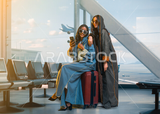 الاستعداد والتجهيز للرحلة الصيفية، بورتريه لرفيقتين عربيتين خليجيتين سعوديتين ترتديان عباءات خليجية عصرية ونظارات شمسية و تجلسان على حقيبة السفر في قاعة الانتظار في المطار، تصوير سيلفي عبر الهاتف المحمول، قضاء أوقات ممتعة مع الأصدقاء في الرحلات السياحية