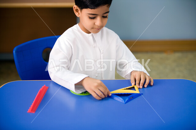 Using geometric shapes and recognizing their forms, a close-up of an Arab Gulf Emirati student wearing the Emirati uniform and holding a compass, learning by playing in the classroom, enjoying recreational activities for preschool students