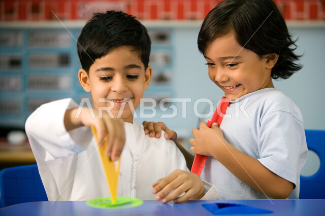 تعلم مهارة التركيز والابتكار لدى الطلاب، صورة مقربة لتلميذين عربيين خليجين اماراتيين يرتديان الزي الكاجوال الاماراتي ويمسكان البوصلة، التعلم عن طريق اللعب داخل الفصل، الاستمتاع بالنشاطات الجماعية الترفيهيه لطلاب التمهيدي