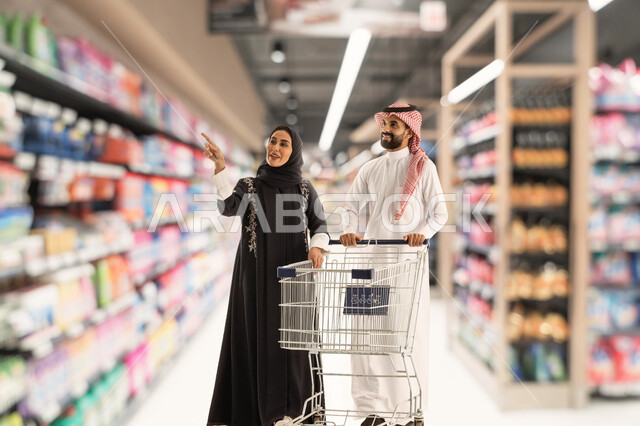 Gestures of joy and happiness in purchasing supplies. A Saudi Gulf Arab man wearing a thobe and shemagh holds a shopping basket in his hand and walks around the store with his wife. A smiling Saudi woman wearing a black abaya joins her husband in buying supplies. Spending a good time in the supermarket.