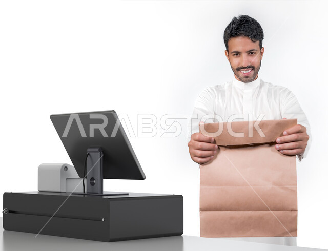 مفهوم الابتسامة بوجه العملاء و التعامل بود معهم, بورتريه لمحاسب عربي خليجي سعودي مبتسم يرتدي الثوب السعودي ويقوم بتسليم كيس التسوق للزبون،  بائع يعمل على ماكينة الكاشير، استلام المشتريات والطلبات عند أمين الصندوق  ، الاستفادة من العروض والخصومات, خلفية بيضاء