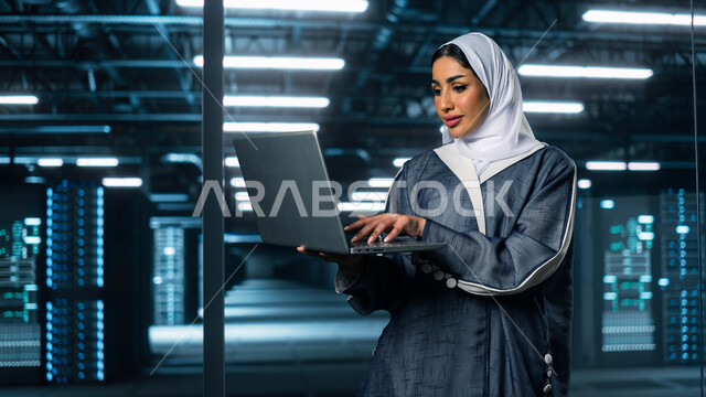 مفهوم  تكنولوجيا التخزين السحابي, إستثمار الخبرات السعودية في بناء مراكز بيانات متطورة وحديثة، صورة مقربة لامرأة عربية خليجية سعودية تستخدم الحاسب المحمول في الإشراف وإدارة معلومات الداتا سنتر, خلفية هولوجرامية ثلاثية الأبعاد لغرفة التحكم بالسيرفرات وخوادم الانترنت