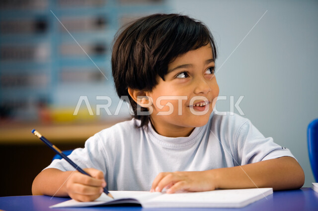 النظر إلى شيء ما بإيماءات السرور , أهداف التربيه والتعليم, صورة مقربة لطالب تمهيدي عربي خليجي إماراتي يرتدي الزي الكاجوال يقوم بكتابة الدرس, الاجتهاد في كتابة التمارين, جودة التعليم في مدارس الإمارات العربية, بداية عام دراسي جديد