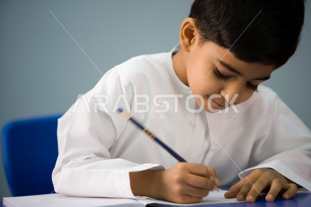 جودة التعليم في مدارس الإمارات العربية, أهداف التربيه والتعليم, صورة مقربة لطالب تمهيدي عربي خليجي إماراتي يرتدي الزي الإماراتي يقوم بكتابة الدرس, الاجتهاد في كتابة التمارين, التركيز والإندماج أثناء الكتابة, بداية عام دراسي جديد