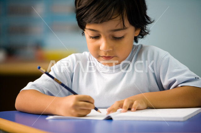 بداية عام دراسي جديد, أهداف التربيه والتعليم, صورة مقربة لطالب تمهيدي عربي خليجي إماراتي يرتدي الزي الكاجوال يقوم بكتابة الدرس, الاجتهاد في كتابة التمارين, التركيز والإندماج أثناء الكتابة, جودة التعليم في مدارس الإمارات العربية