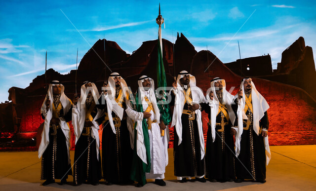 Standing in front of Salwa Palace in Diriyah, traditional national events, the Hijazi Ardha band looking at something, Saudi popular folklore, wearing the traditional dhokla and bralette, Saudi Flag Day, March 11, celebrating the anniversary of the National Day, September 23, the concept of love for the homeland and belonging to it.