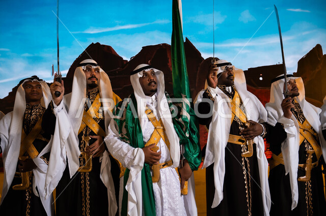 Standing in front of Salwa Palace in Diriyah, traditional national occasions, the Hijazi Ardha band looking at something, Saudi popular folklore, wearing the traditional dhokla and bra, Saudi Flag Day, March 11, celebrating the anniversary of the National Day, Rabi’ al-Awwal 8, the concept of love for the homeland and belonging to it.