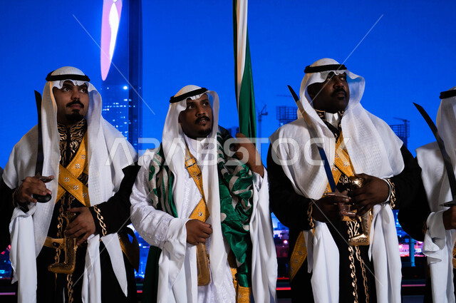 Looking at something, a close-up of the Hijazi Ardha band wearing the popular Dakla and Vermeela costume, popular events on national occasions, Eid and the Saudi National Day on September 23, celebrating the Saudi Flag Day on March 11, the background of the Kingdom Tower and skyscrapers in Riyadh