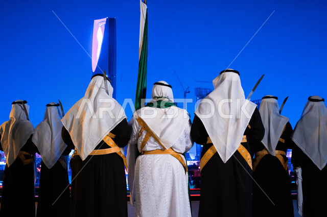 الانشطة والفعاليات الوطنية الشعبية في المناسبات، اليوم الوطني السعودي في 23 سبتمبر، صورة مقربة من الخلف لفرقة العرضة الحجازية ترقص بالسيوف ترتدي زي الدقلة والفرميلة الشعبية، الاحتفال بيوم العلم السعودي 11 مارس، ابراج وناطحات سحاب مدينة الرياض