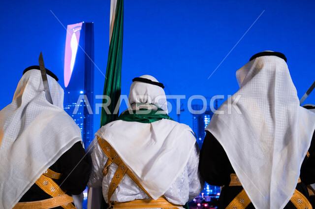  الفعاليات الوطنية الشعبية في المناسبات، ذكرى اليوم الوطني السعودي في 23 سبتمبر، صورة مقربة من الخلف لفرقة العرضة الحجازية ترقص بالسيوف ترتدي زي الدقلة والفرميلة الشعبية، الاحتفال بيوم العلم السعودي 11 مارس، خلفية برج المملكة وناطحات السحاب