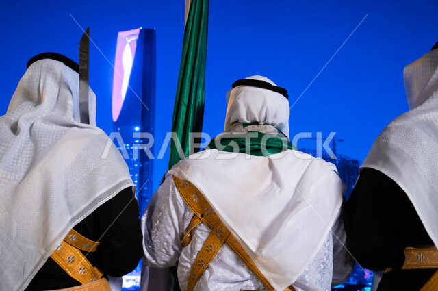  الفعاليات الوطنية الشعبية في المناسبات، اليوم الوطني السعودي في 23 سبتمبر، صورة مقربة من الخلف لفرقة العرضة الحجازية ترقص بالسيوف ترتدي زي الدقلة والفرميلة الشعبية، الاحتفال بيوم العلم السعودي 11 مارس، خلفية برج المملكة وناطحات السحاب