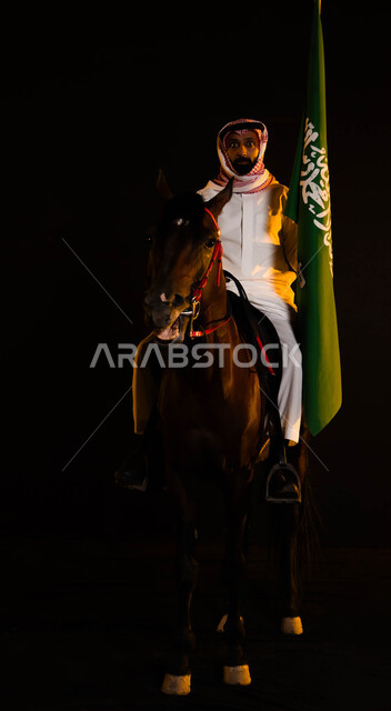 إظهار التفاني والانتماء للبلاد، ذكرى يوم العلم السعودي 11 مارس، اتباع تقاليد الأجداد بتربية الخيول، العيد الوطني السعودي 23 سبتمبر، الاحتفال باليوم الوطني، شاب عربي سعودي يرتدي الشماغ والزي التقليدي يحمل علم السعودية يمتطي حصانه الاصيل