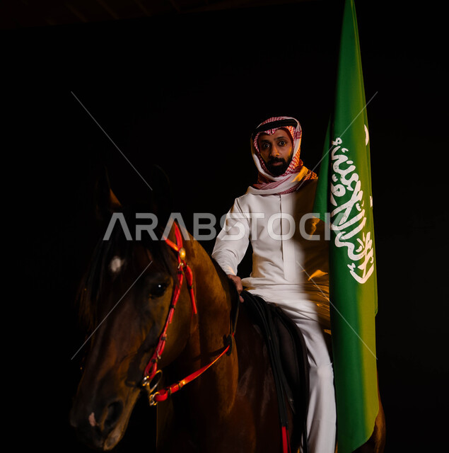 اتباع تقاليد الأجداد بتربية الخيول، حب الوطن والانتماء إليه، ذكرى يوم العلم السعودي 11 مارس، العيد الوطني السعودي 8 ربيع الأول، الاحتفال باليوم الوطني، صورة مقربة لشاب عربي سعودي خليجي يرتدي الشماغ والزي التقليدي يحمل علم السعودية يمتطي حصانه الاصيل