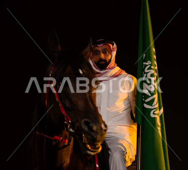 حب الوطن والانتماء إليه، ذكرى يوم العلم السعودي 11 مارس، اتباع تقاليد الأجداد بتربية الخيول، العيد الوطني السعودي 8 ربيع الأول، الاحتفال باليوم الوطني، صورة مقربة لشاب عربي سعودي خليجي يرتدي الشماغ والزي التقليدي يمسك علم السعودية يمتطي حصانه الاصيل