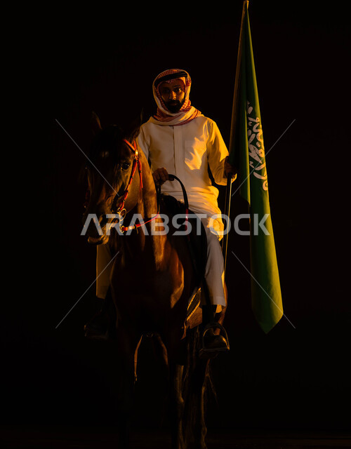 إظهار التفاني والانتماء للبلاد، ذكرى يوم العلم 11 مارس، اتباع تقاليد الأجداد بتربية الخيول، العيد الوطني السعودي 23 سبتمبر، الاحتفال في اليوم الوطني السعودي، شاب عربي سعودي يرتدي الشماغ والزي التقليدي يحمل علم السعودية يمتطي حصانه الاصيل