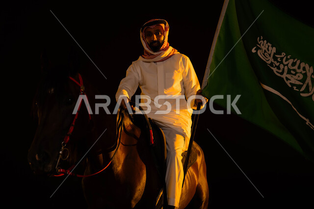 إظهار الولاء للمملكة وتعزيز الروح الوطنية، ذكرى يوم العلم السعودي 11 مارس، اتباع تقاليد الأجداد بتربية الخيول، العيد الوطني 8 ربيع الأول، الاحتفال في اليوم الوطني السعودي، شاب عربي سعودي يرتدي الشماغ والزي التقليدي يحمل علم السعودية يمتطي حصانه الاصيل