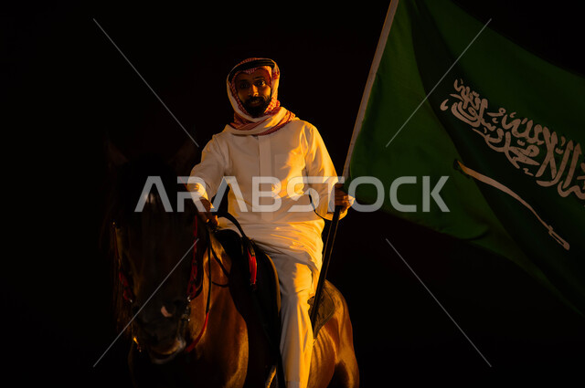 التباهي والفخر في الهوية الوطنية، ذكرى يوم العلم 11 مارس، اتباع تقاليد الأجداد بتربية الخيول، العيد الوطني السعودي 8 ربيع الأول، الاحتفال باليوم الوطني السعودي، شاب عربي سعودي خليجي يرتدي الشماغ والزي التقليدي يمسك علم السعودية يمتطي حصانه الاصيل