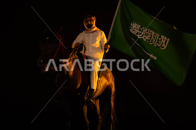 اتباع تقاليد الأجداد في تربية الخيول، التعبير عن الحب والولاء للمملكة، ذكرى يوم العلم السعودي 11 مارس، العيد الوطني 23 سبتمبر، الاحتفال في اليوم الوطني السعودي، شاب عربي سعودي خليجي يرتدي الشماغ والزي التقليدي يمسك علم السعودية يمتطي حصانه الاصيل