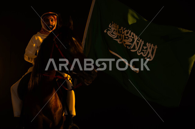 التعبير عن الحب والولاء للمملكة، ذكرى يوم العلم السعودي 11 مارس، اتباع تقاليد الأجداد بتربية الخيول، العيد الوطني 23 سبتمبر، الاحتفال باليوم الوطني السعودي، شاب عربي سعودي خليجي يرتدي الشماغ والزي التقليدي يمسك علم السعودية يمتطي حصانه الاصيل