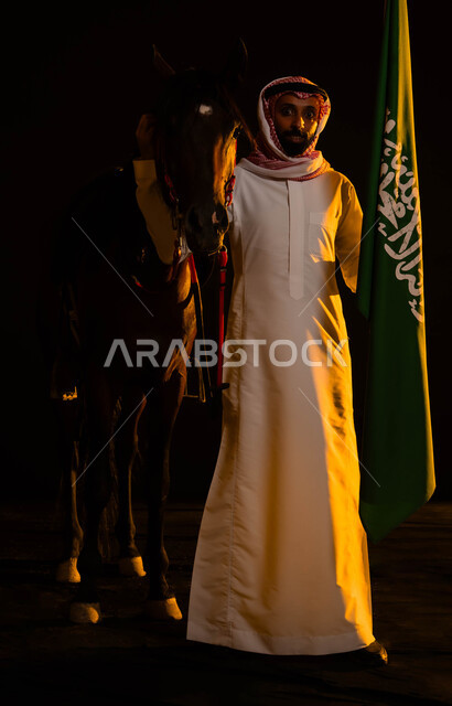 العيد الوطني السعودي 23 سبتمبر، ذكرى يوم العلم 11 مارس، الوقوف بفخر واعتزاز بالوطن أمام الفرس والنظر إلى الكاميرا، صورة مقربة لرجل عربي سعودي يرتدي الشماغ والثوب السعودي يمسك علم السعودية يحتفل باليوم الوطني السعودي، صورة الجسم بالطول الكامل