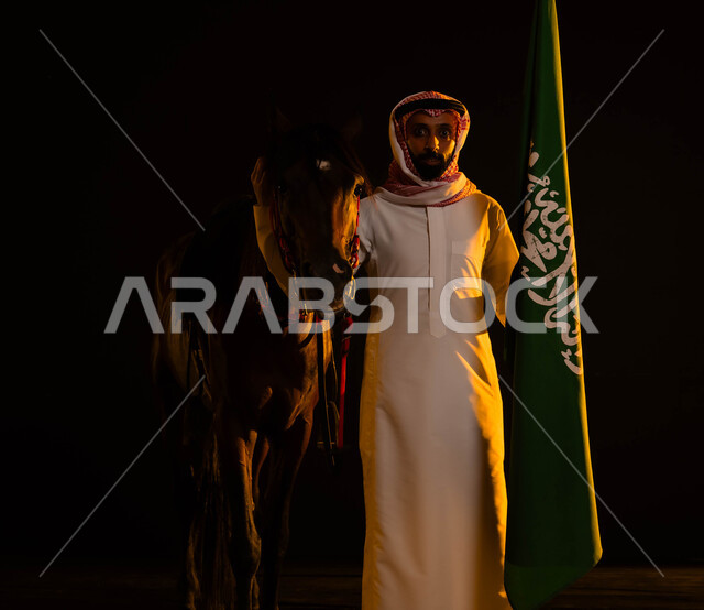 استذكار التاريخ و الهوية الوطنية، العيد الوطني السعودي 23 سبتمبر، ذكرى يوم العلم 11 مارس، الوقوف بفخر واعتزاز بالوطن أمام الفرس و النظر إلى الكاميرا، صورة مقربة لرجل عربي سعودي يرتدي الشماغ والثوب السعودي يمسك علم السعودية يحتفل باليوم الوطني السعودي