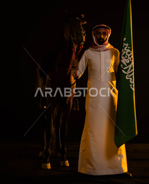 الوقوف بفخر واعتزاز بالوطن أمام الفرس و النظر إلى الكاميرا، ذكرى يوم العلم السعودي 11 مارس، العيد الوطني 8 ربيع الأول، صورة مقربة لرجل عربي سعودي يرتدي الشماغ والثوب السعودي يمسك علم السعودية يحتفل باليوم الوطني السعودي، صورة الجسم بالطول الكامل
