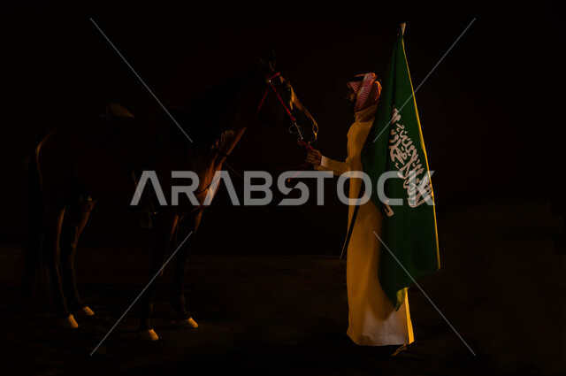 المناسبات و الأعياد الوطنية، نشر حب الخيل بين ابناء الوطن، العيد الوطني السعودي 23 سبتمبر، ذكرى يوم العلم السعودي 11 مارس، الاحتفال في اليوم الوطني، صورة من الجانب لشاب عربي سعودي يرتدي الشماغ والثوب السعودي يمسك علم السعودية يقوم بملاعبة فرسه