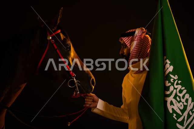 الأعياد و المناسبات الوطنية، نشر حب الخيل بين ابناء الوطن، العيد الوطني السعودي 23 سبتمبر، ذكرى يوم العلم السعودي 11 مارس، الاحتفال في اليوم الوطني، صورة مقربة لشاب عربي سعودي يرتدي الشماغ والثوب السعودي يمسك علم السعودية يقوم بملاعبة فرسه في الليل