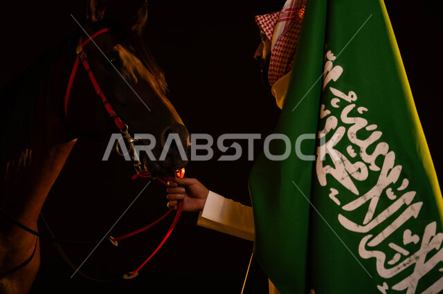 ذكرى يوم العلم السعودي 11 مارس، الاحتفال مع الخيول في اليوم الوطني، العيد الوطني السعودي 8 ربيع الاول، المناسبات والأعياد الوطنية، صورة مقربة من الجانب لشاب عربي سعودي يرتدي الشماغ والزي التقليدي يمسك علم السعودية يقوم بملاعبة فرسه في الليل