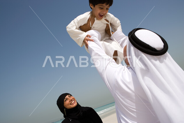 Spending an enjoyable summer vacation, family leisure activities on vacation, a pleasant family atmosphere, a close-up of an Arab Emirati father playing with his son and raising him high in the air, enjoying summer activities, beach resorts in the Emirate