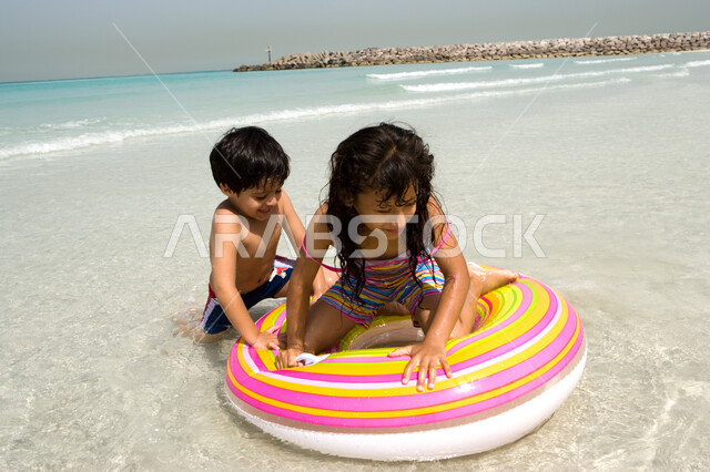 أخوان يستمتعان باللعب في أجواء صيفية ممتعة, فتاة صغيرة عربية إمراتية خليجية تجلس داخل عوامة السباحة على شاطئ البحر وتقوم باللعب رفقة أخيها في رحلة صيفية, أنشطة ترفيهية عائلية في العطلة, الإستمتاع بالفعاليات مع الأخوة, منتجعات شاطئية في الإمارات