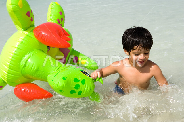 قضاء أوقات صيفية ممتعة بالقيام بنشاطات ترفيهية على البحر , الاستمتاع باللعب في الساحل, صبي عربي خليجي إماراتي يمسك بيده عوامة السباحة ويسبح داخل البحر في إجازة الصيف، السياحة في دول الخليج العربي