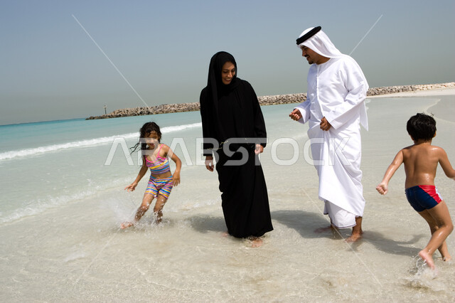 التسلية و قضاء أوقات الإجازة مع الأبناء, أخوان يلعبان لعبة المطاردة ويجريان حول والديهم, السياحة في دول الخليج العربي والاستمتاع بالعطلة الصيفية، مفهوم الاسرة الإماراتية السعيدة، أب وأم عربيان خليجيان إماراتيان يقومان باللعب مع أبنائهم على شاطئ البحر