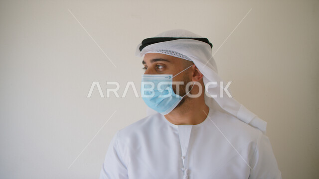 ايماءات شرود الذهن والنظر إلى شيء ما، إتباع الاحترازات و التعليمات الوقائيه ضد الفايروسات، تجنب الملامسه السطحيه والحفاظ على المسافات، بورتريه مقرب لرجل عربي خليجي اماراتي يرتدي الكندوره والغتره يضع القناع الطبي للوقايه من فايروس كورونا، خلفيه بيضاء.